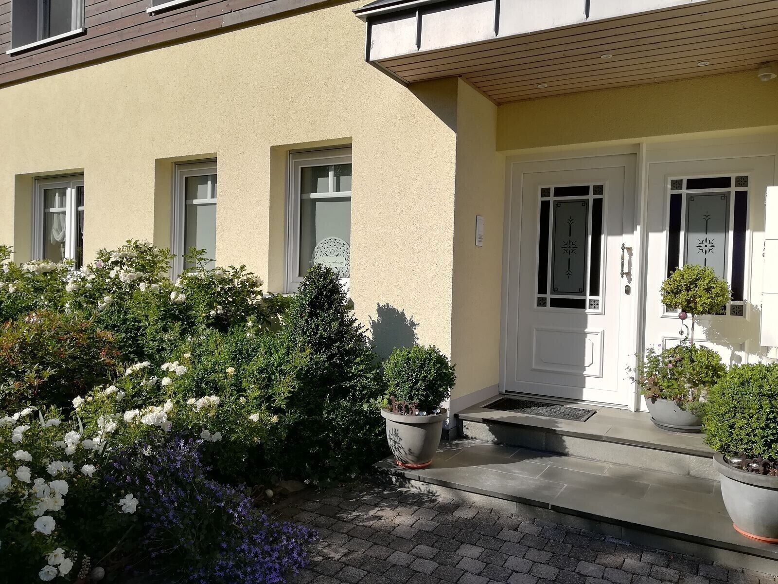 A building with a beige facade, white doors, and windows. There are potted plants and a paved entrance area.