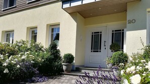 A building with a number 20 sign, a white door, and a paved entrance area with lavender and white flowers. - Attic Apartment (Olsberg)