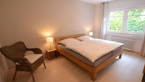 A bedroom with a wooden bed, a wicker chair, a nightstand with a lamp, and a window with a view of greenery. - Apartment with 2 Bedrooms, Sleeps up to 4 Guests (Olsberg)