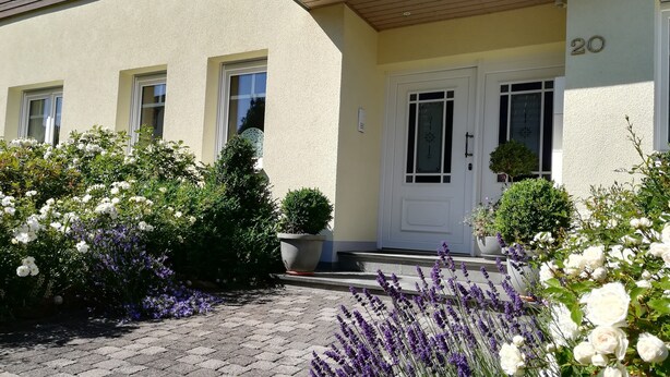 A building with a beige facade, a white door with a number plaque, and a paved entrance area with lavender and white flowers. - Apartment with 2 Bedrooms, Sleeps up to 4 Guests (Olsberg)