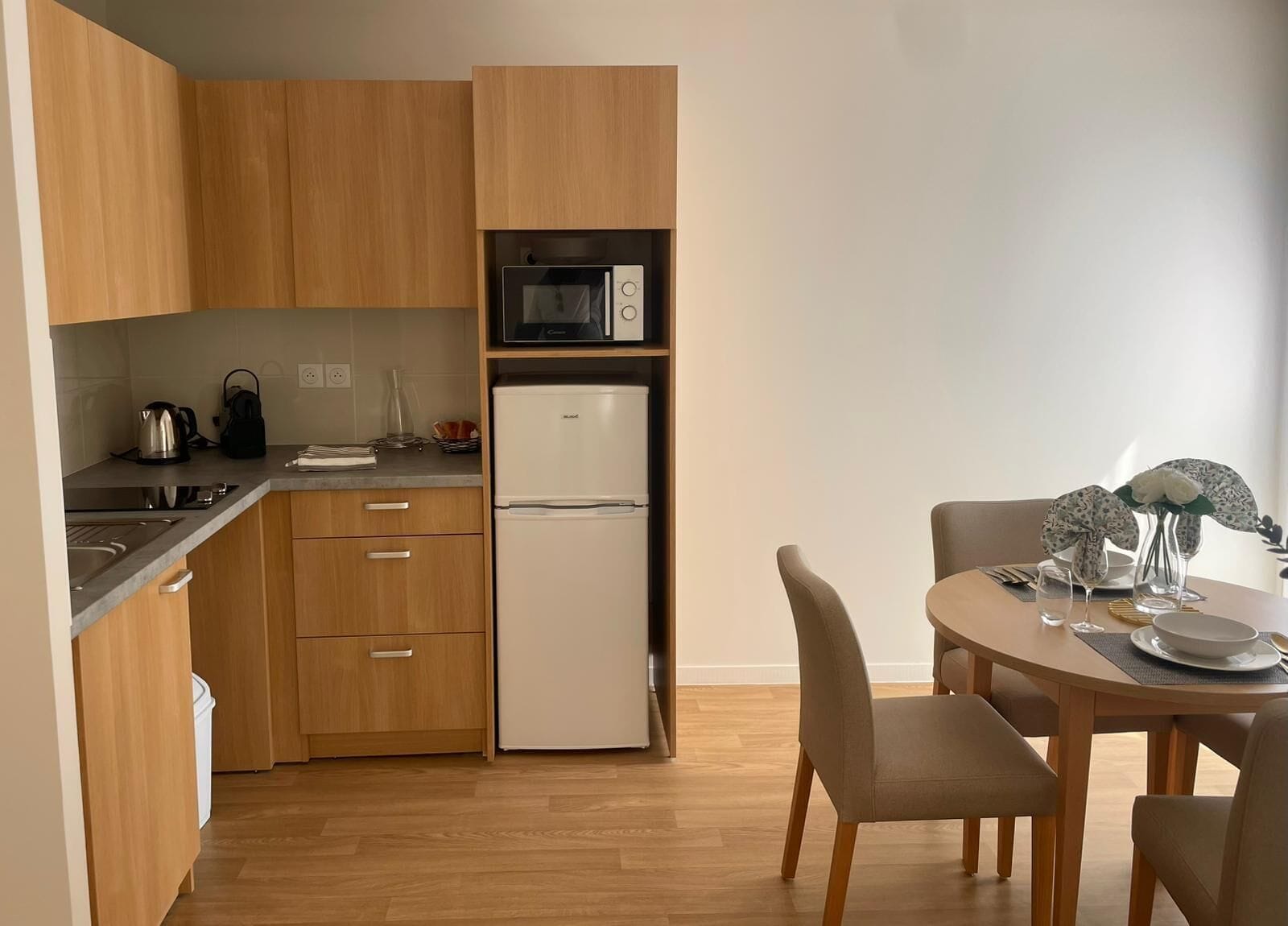 Una cucina moderna con mobili in legno, forno a microonde integrato e frigorifero bianco. Si nota un tavolo da pranzo con sedie e un vaso di fiori.