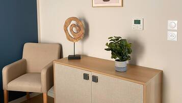 A room with a beige armchair, a wooden cabinet with two doors, a potted plant, a wall-mounted thermostat, and a framed artwork above the cabinet.