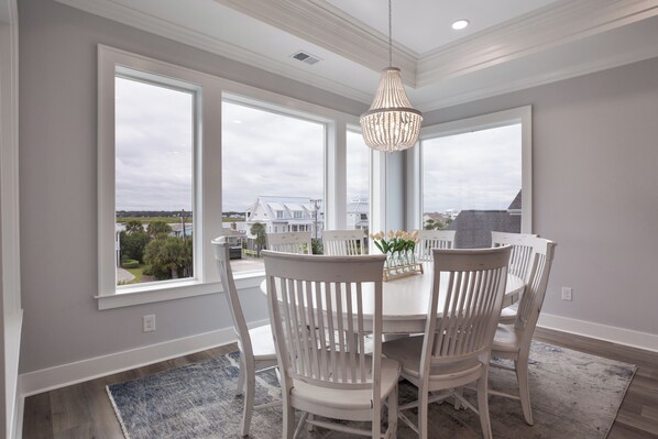 A dining area with a white table and chairs, large windows, and a chandelier.