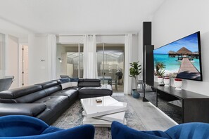 A modern living room with a black leather sofa, a white coffee table, and a flat-screen TV mounted on the wall displaying a beach scene.