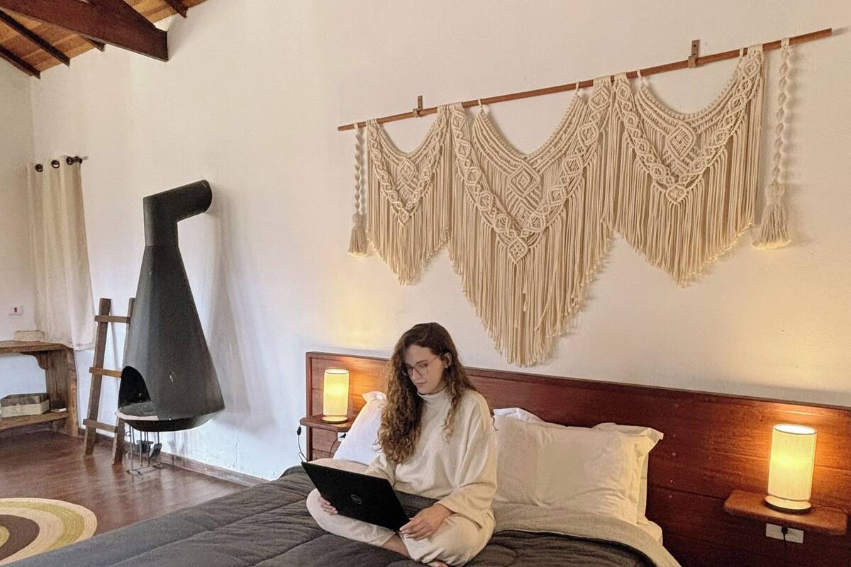 A bed with a dark grey bedspread, wooden headboard, and a macrame wall hanging. There is a small wooden table with a lamp beside the bed, and a black conical heater on the floor.
