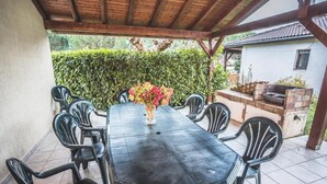 A covered patio with a table and chairs, a grill area, and a white building in the background. - Gîtes chez PAT, a house in the countryside near the beach (Ychoux)