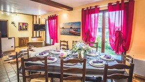 A dining area with a table set for six, red curtains, a painting of a boat on the wall, and a TV in the corner. - Gîtes chez PAT, a house in the countryside near the beach (Ychoux)