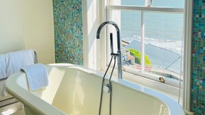 A bathroom with a freestanding bathtub, a window with a view of the ocean, and a tiled wall. - The Hambrough (Ventnor)
