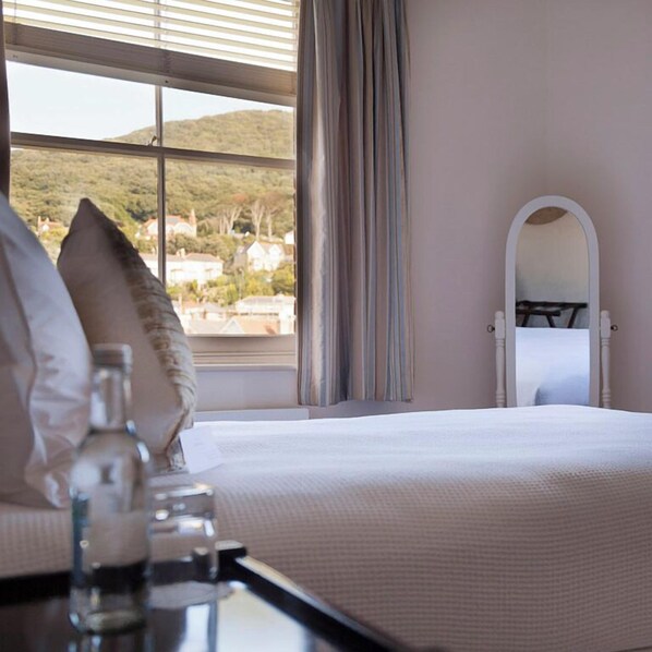 A bedroom with a large window, a bed, a bedside table with a glass bottle, and a mirror on the wall. - The Hambrough (Ventnor)