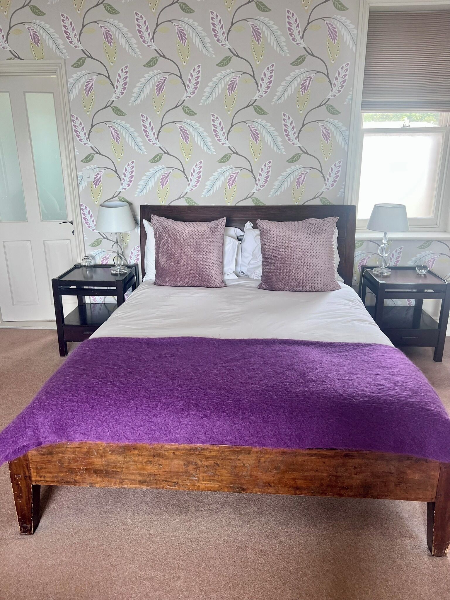 A bedroom with a large bed, two nightstands, a window with blinds, and a floral wallpaper pattern.