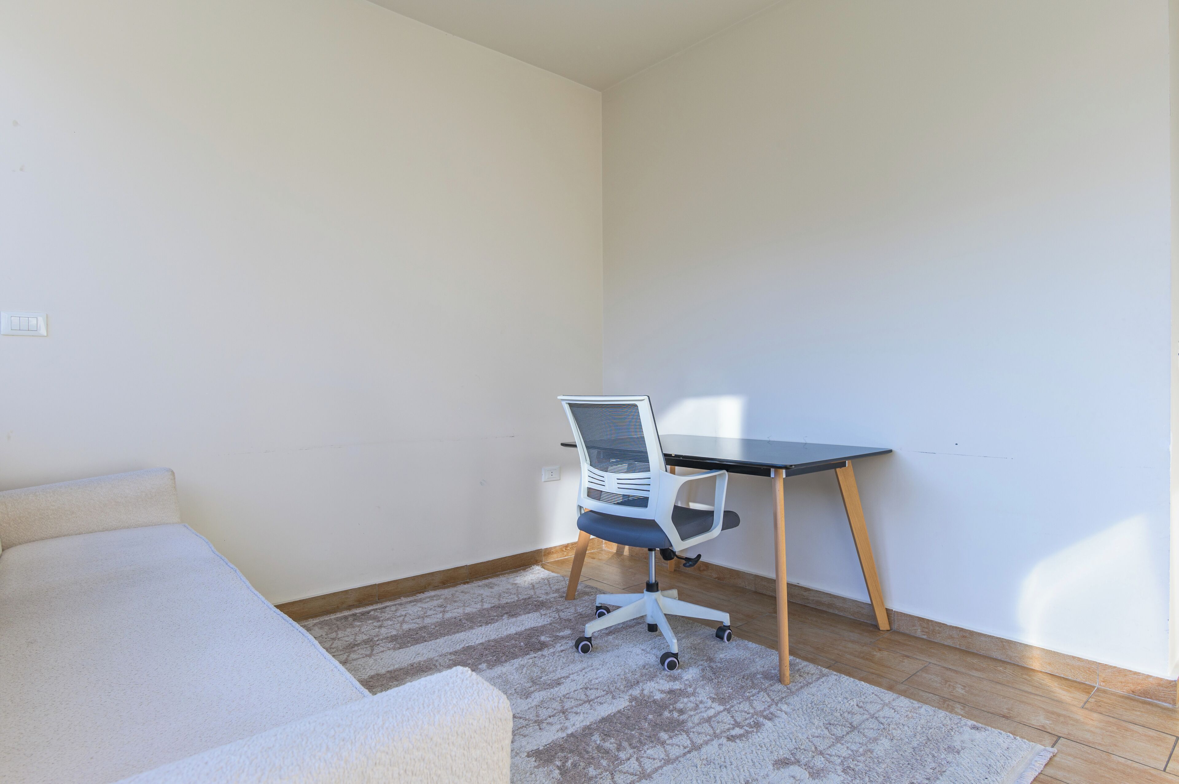 Una habitación pequeña y poco amueblada, con un sofá blanco, un escritorio con una silla y una alfombra estampada.