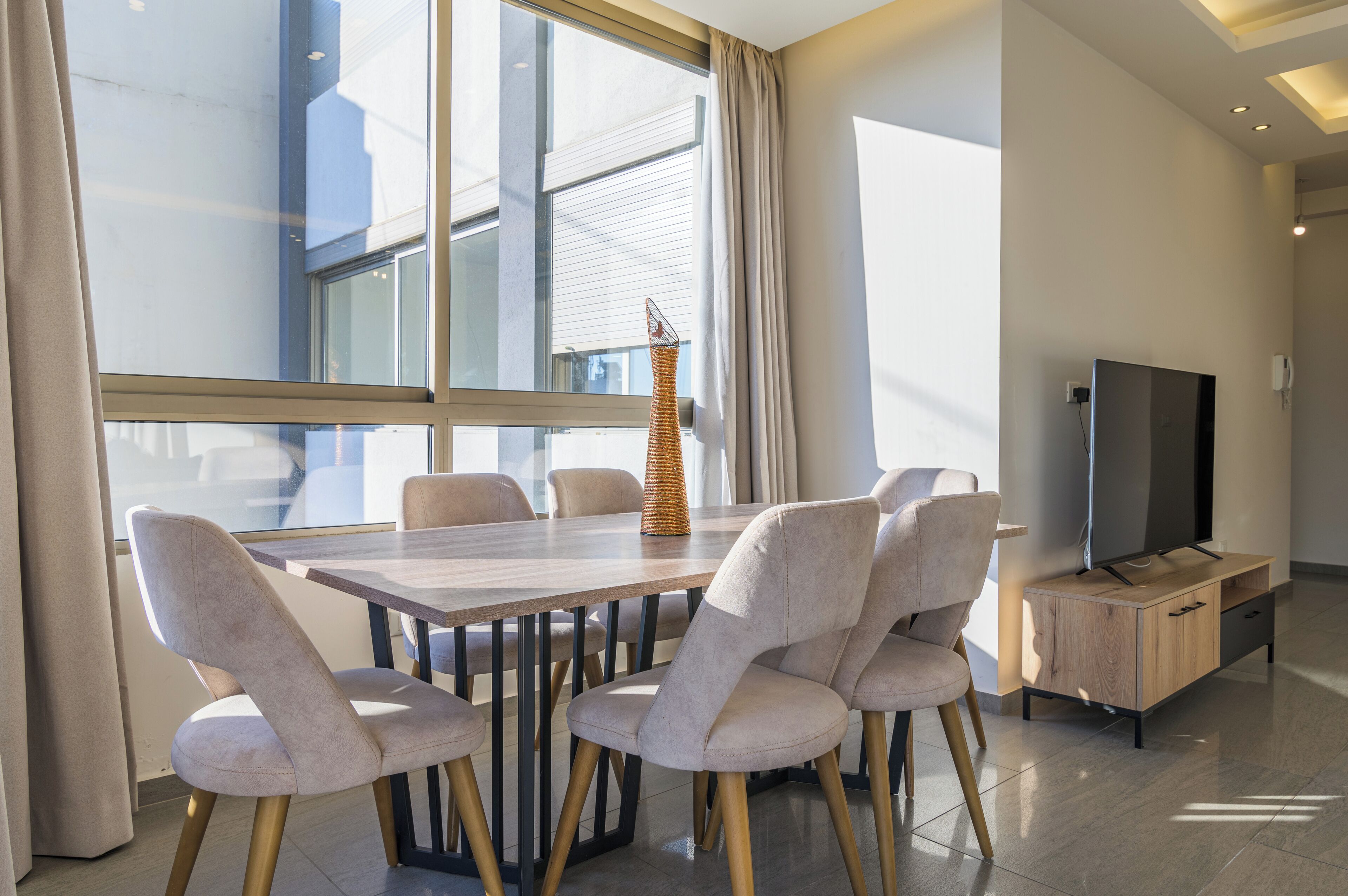 A modern dining area with a wooden table and six chairs, a tall vase, and a television on a wooden cabinet.