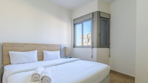 A neatly made bed with white linens and rolled towels, a wooden headboard, and a window with a view of buildings outside. - Elevate - Sleek 2-BR Apt by Santa Maria, Jal el Dib (Jal El Dib)