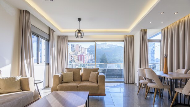 A modern living room with a sofa, a dining table with chairs, and large windows offering a city view. - Elevate - Sleek 2-BR Apt by Santa Maria, Jal el Dib (Jal El Dib)