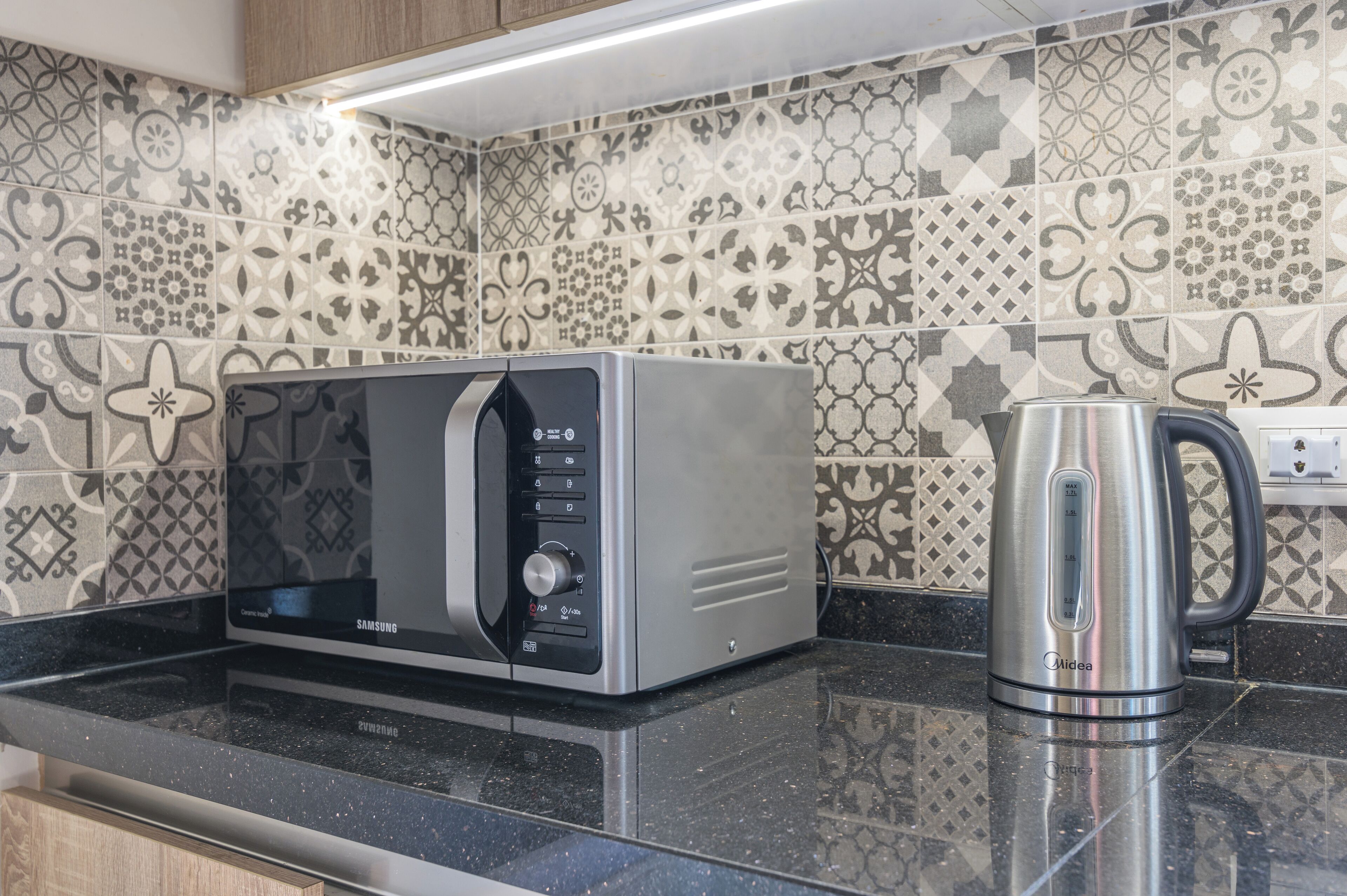 A kitchen with a stainless steel microwave, a kettle, and patterned tiles on the backsplash.