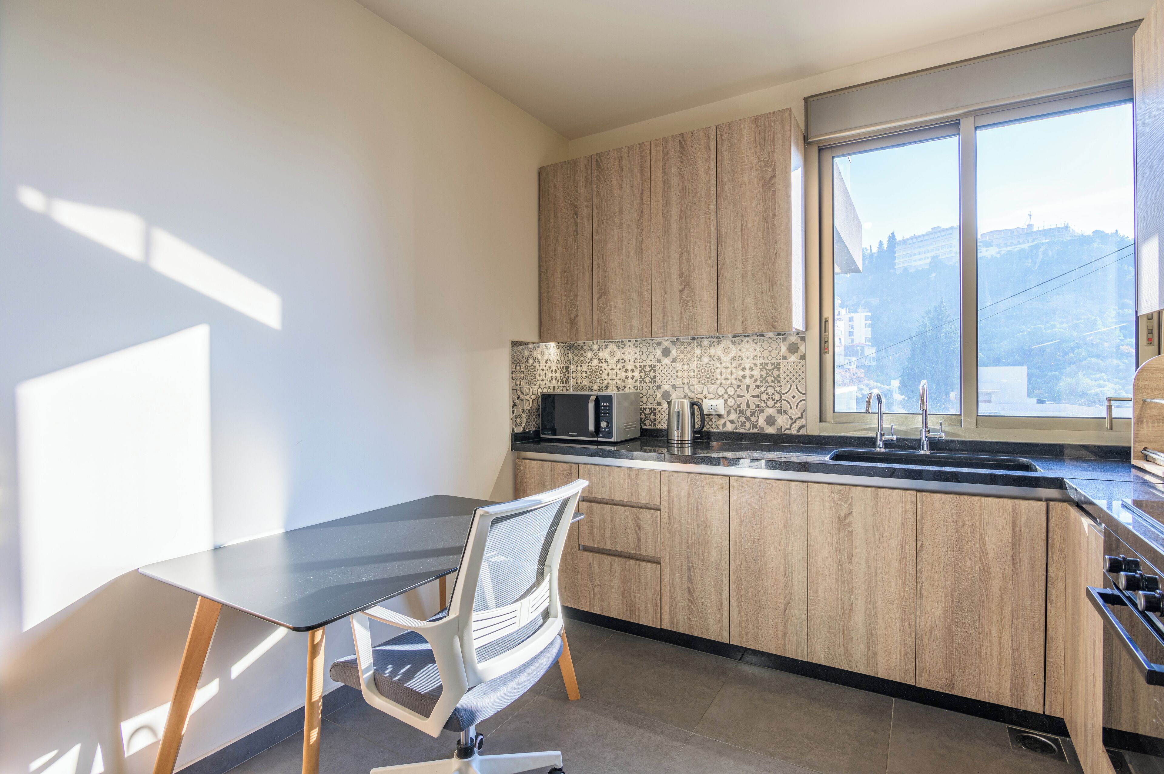 A modern kitchen with wooden cabinets, a black countertop, a microwave, and a large window with a view of a hill.