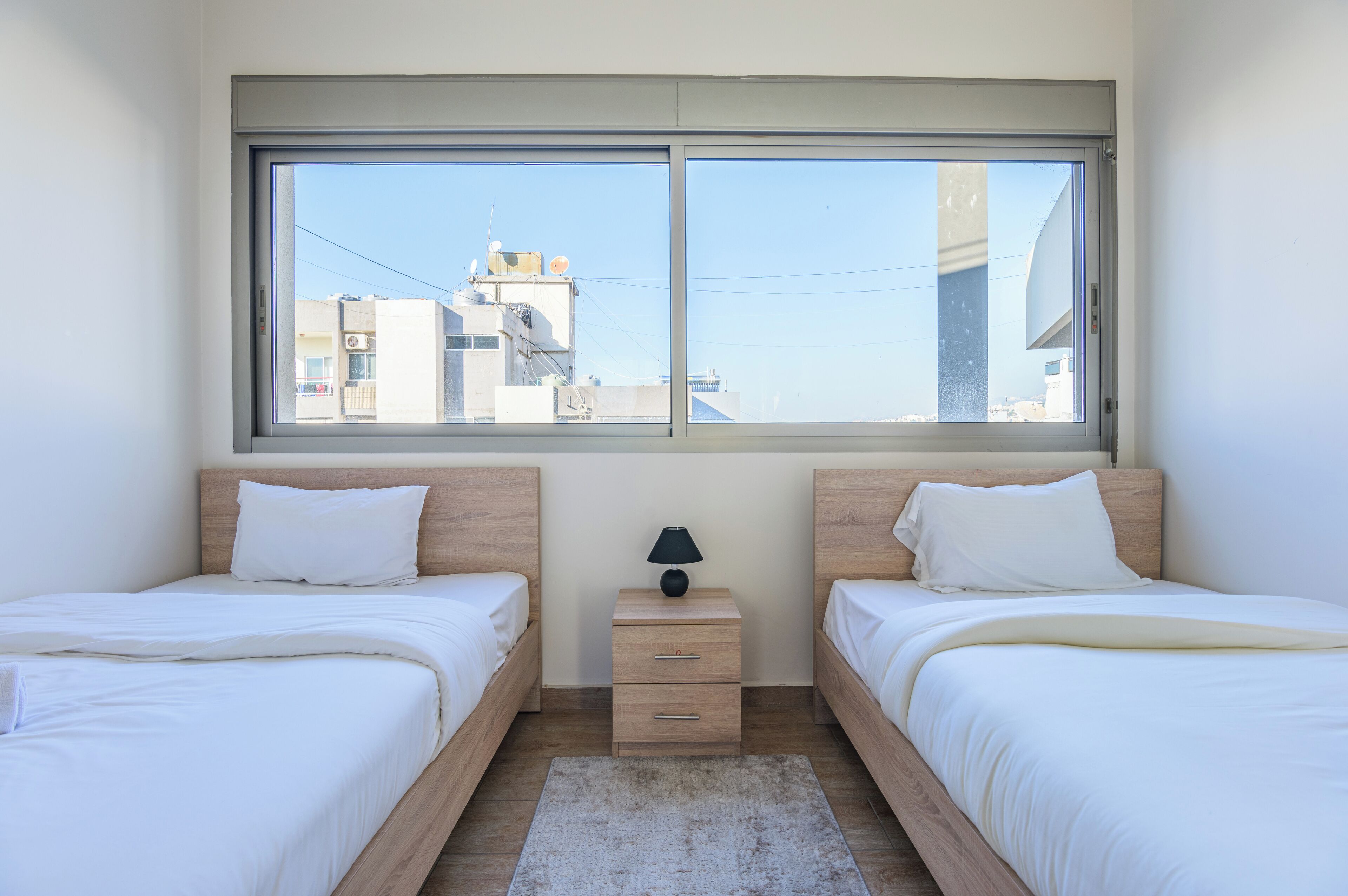 A bedroom with two beds, a nightstand with a lamp, and a window with a view of buildings.