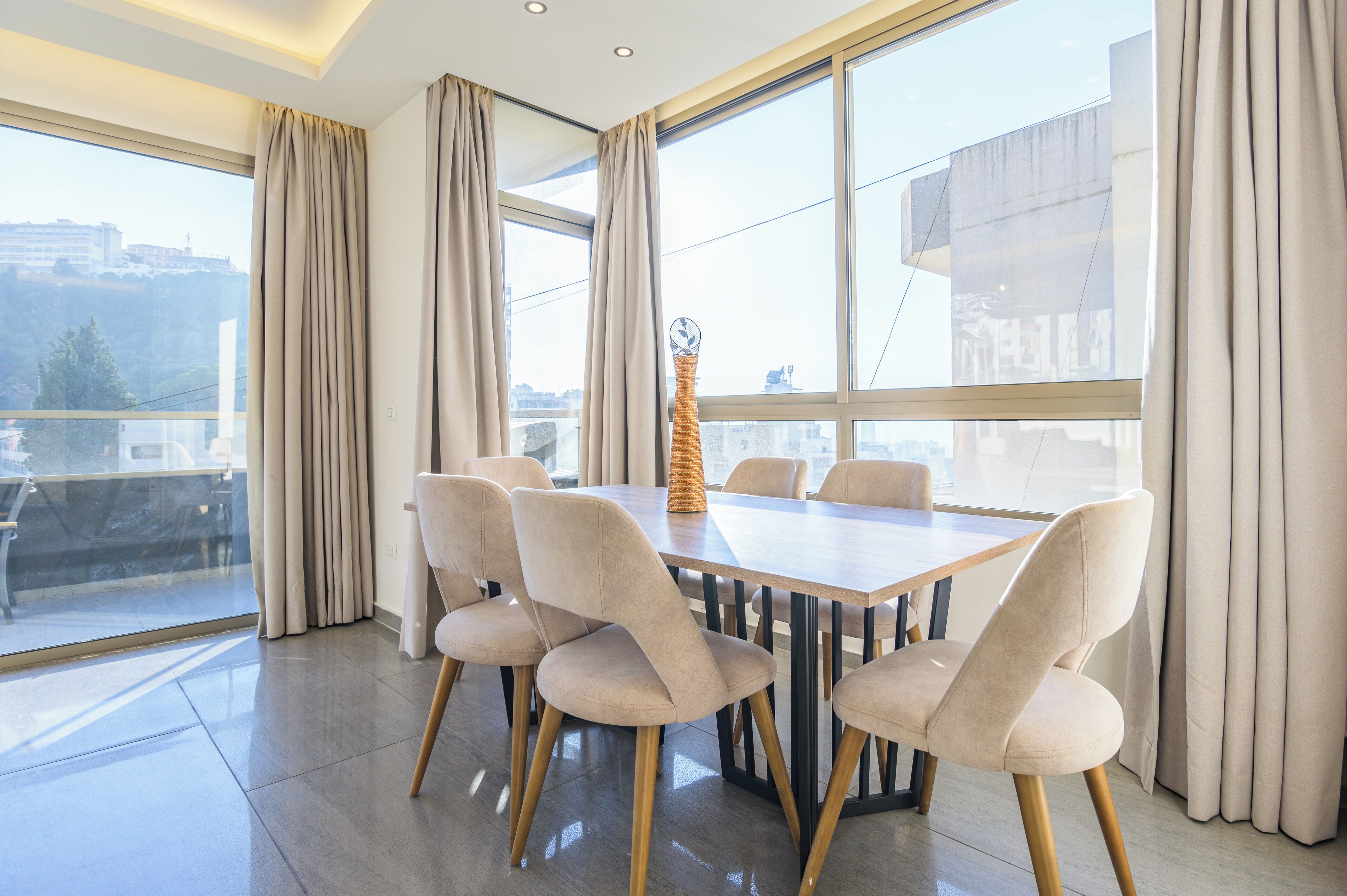 A modern dining area with a rectangular table and six chairs, large windows with curtains, and a view of the cityscape.
