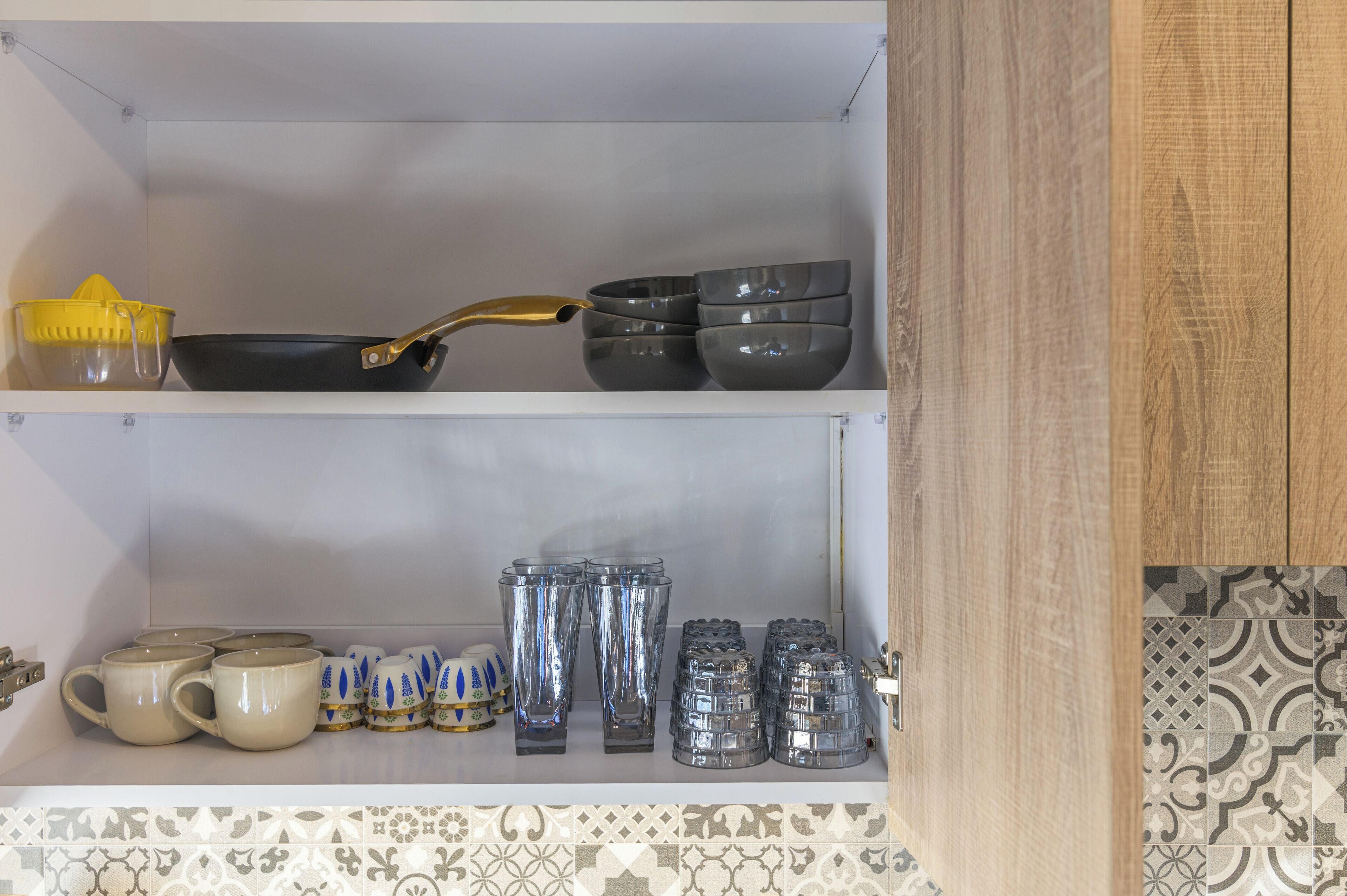 A kitchen shelf with various kitchenware including a yellow colander, a black frying pan, and several bowls and mugs.