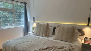 Ein Schlafzimmer mit einem großen Bett, zwei Nachttischen, einem Fenster mit Vorhängen und Blick auf Bäume.