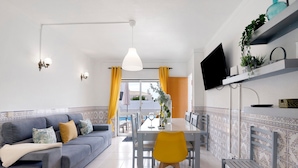 Un salon moderne avec un canapé, une table à manger et des chaises, un téléviseur fixé au mur et un balcon agrémenté de rideaux jaunes.