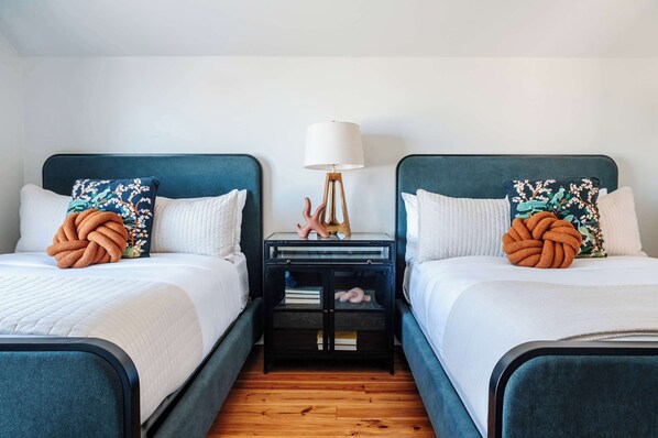 A hotel room with two beds, each with a blue headboard, white bedding, and decorative pillows. There is a nightstand with a lamp and a small shelf between the beds.