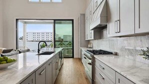 A modern kitchen with a large island, stainless steel appliances, and a view of a cityscape through a sliding glass door. - The 1300 | 4 Home Combo, 28 bedrooms | Heated Pool (New Orleans)