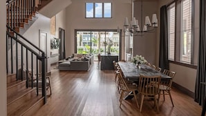 A spacious living area with a wooden staircase, a dining table with chairs, and a large window providing a view of the outdoors. - The 1300 | 4 Home Combo, 28 bedrooms | Heated Pool (New Orleans)