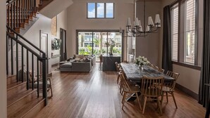 A spacious living area with a wooden staircase, a dining table with chairs, and a large window providing a view of the outdoors. - The 1300 | 4 Home Combo, 28 bedrooms | Heated Pool (New Orleans)