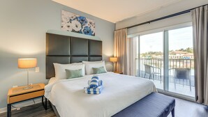 Une chambre d’hôtel avec un grand lit, une banquette, une table de chevet, une fenêtre donnant sur la ville et un tableau au mur.