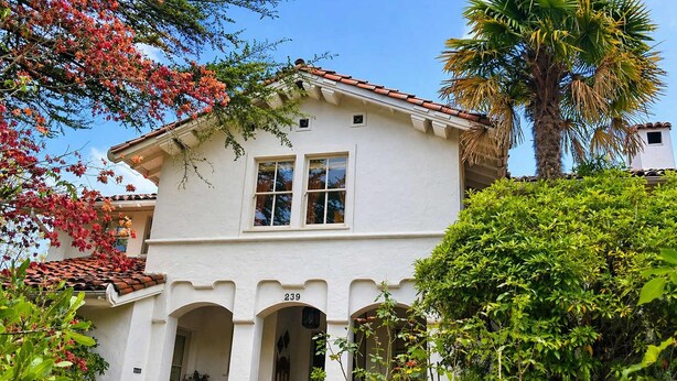 A white two-story house with a red-tiled roof, a number '239' on the front, and a covered porch with potted plants.