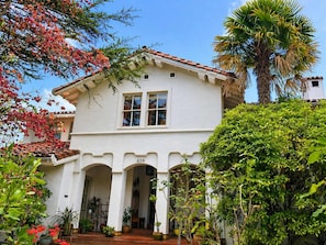 A white two-story house with a red-tiled roof, a number '239' on the front, and a covered porch with potted plants.