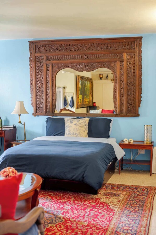 A bedroom with a large ornate mirror above a bed, a red chair, a nightstand with a lamp, and a patterned rug.