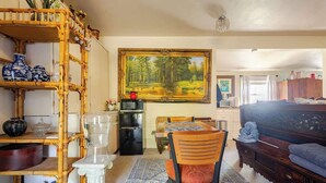 A room with a dining table, chairs, a sofa, a painting, a bookshelf, and a lamp.