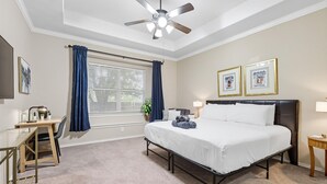 Ein Hotelzimmer mit Bett, Schreibtisch, Stuhl, Deckenventilator, Fenster mit Vorhängen und zwei gerahmten Bildern an der Wand.