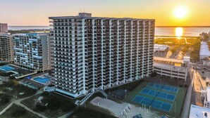 Un edificio de gran altura con varios balcones, piscina, canchas de tenis y vista del atardecer sobre el agua.