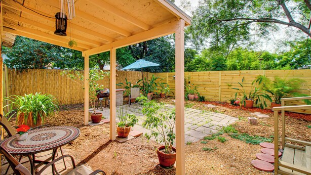 A covered patio area with a wooden ceiling, a round table with chairs, potted plants, and a small garden with stepping stones.