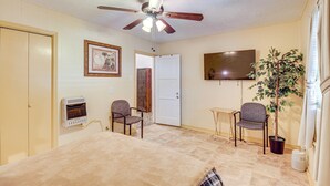 A hotel room with a bed, two chairs, a ceiling fan, a TV, a plant, and a door leading to another room.