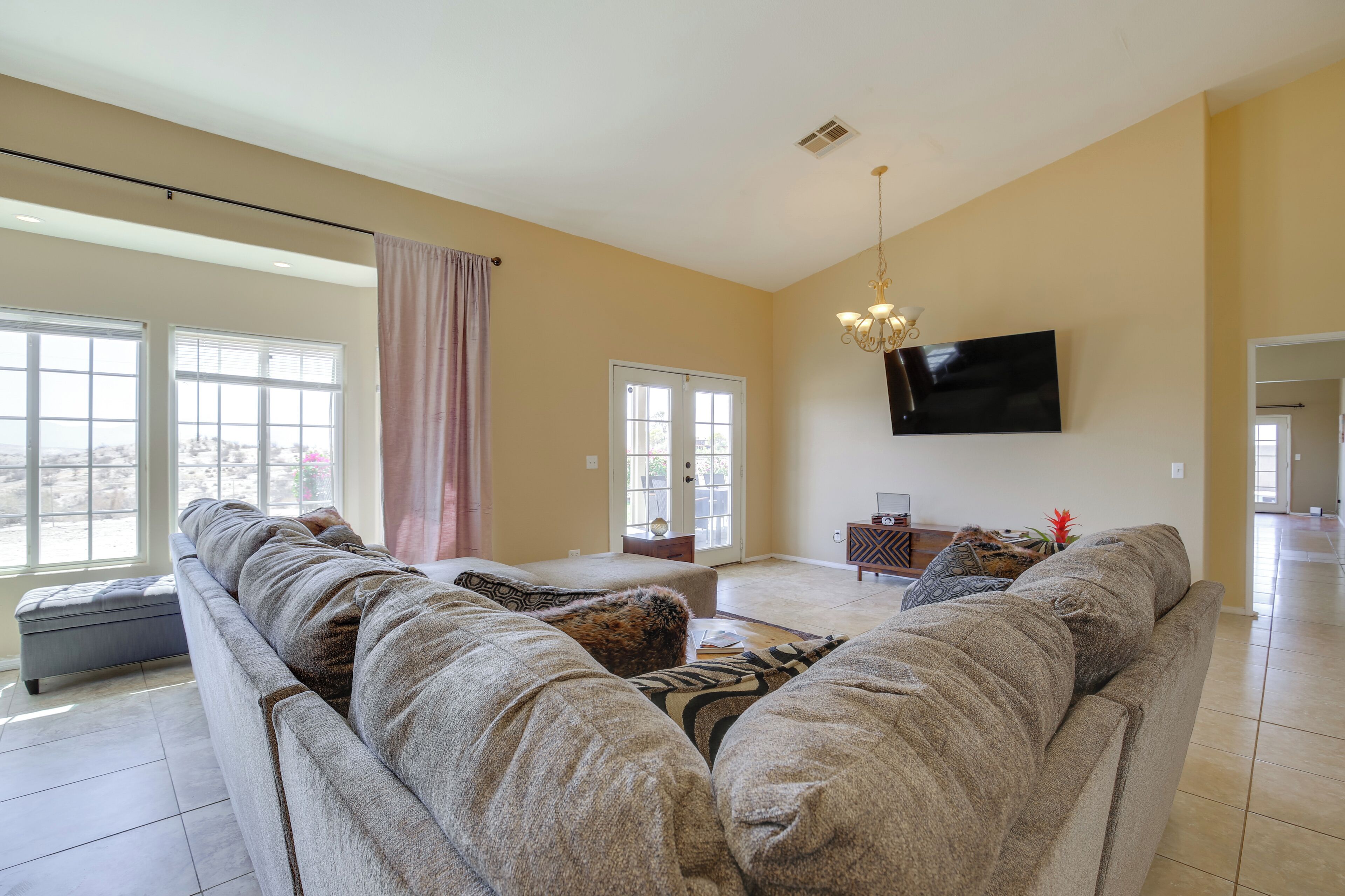 A spacious living room with a large sofa, a flat-screen TV mounted on the wall, and a chandelier.
