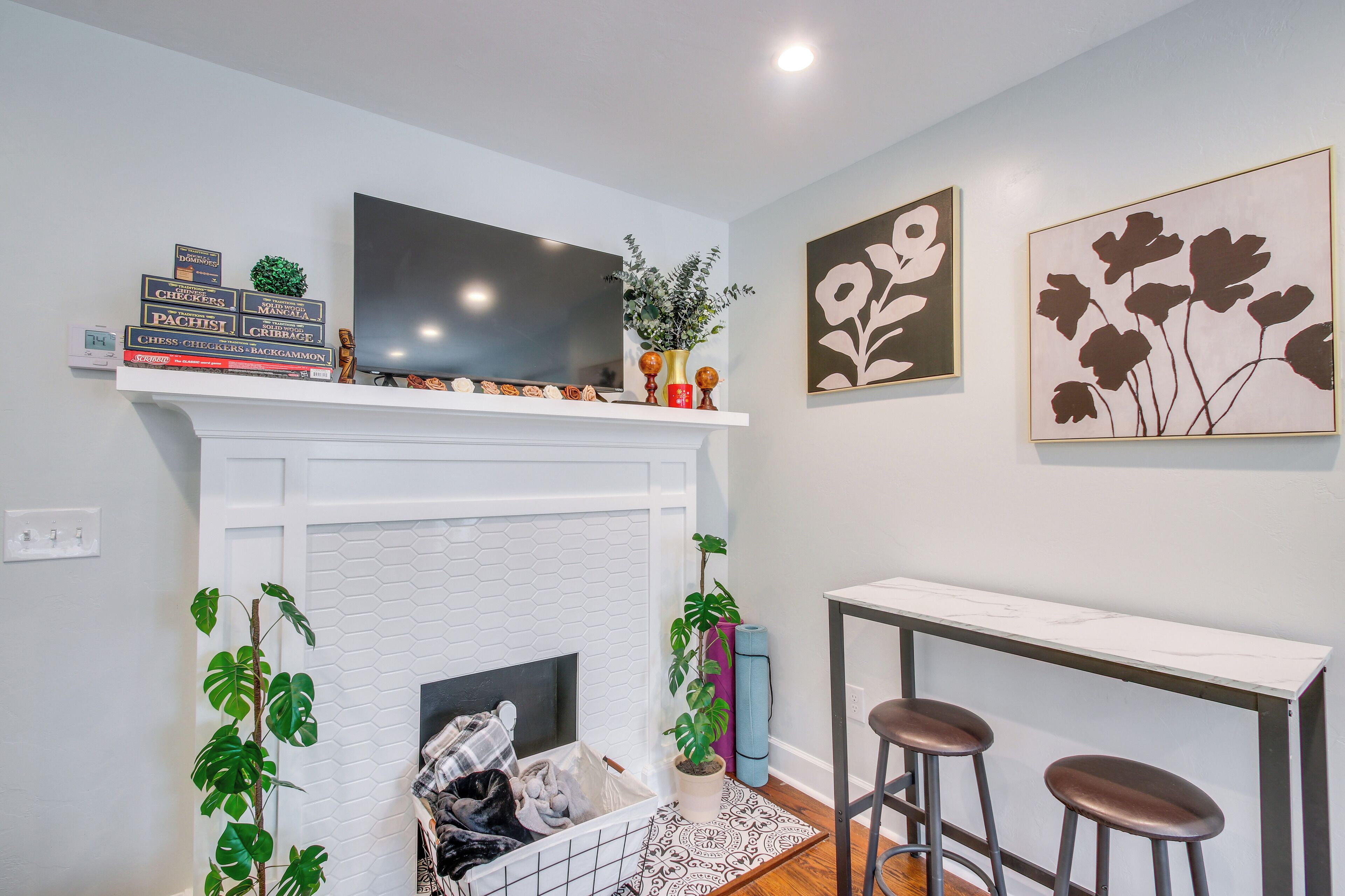 A living room with a white fireplace, a mounted TV, a bookshelf with chess books, and two framed floral paintings.
