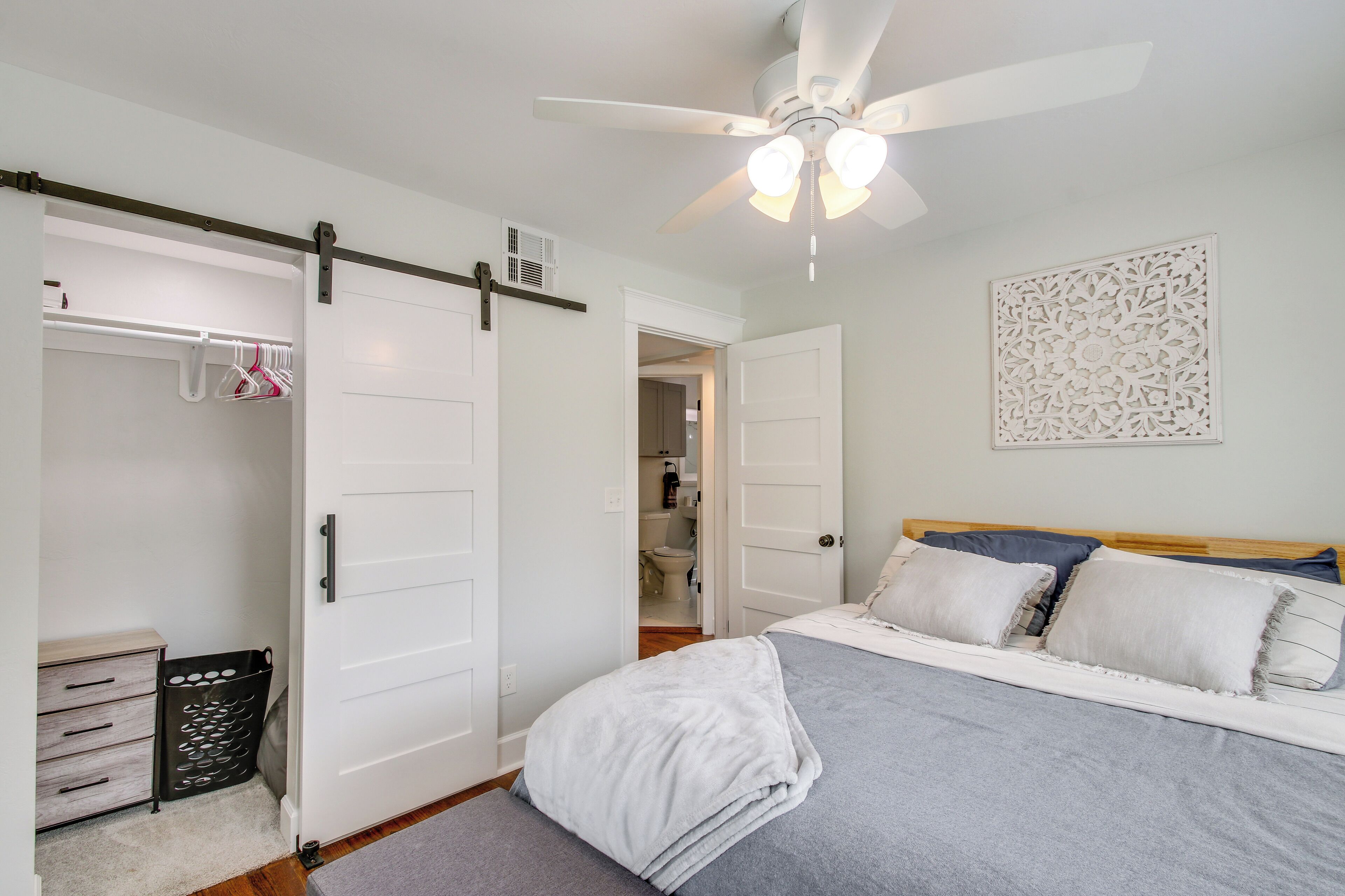 Ein Schlafzimmer mit Bett, Deckenventilator, Kleiderschrank mit Kleidung und einem durchgegangenen Tür sichtbaren Badezimmer.