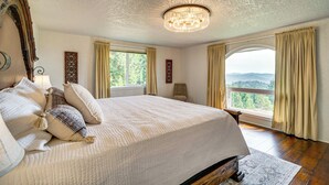 A bedroom with a large bed, a chandelier, a view of the outdoors, and a wooden floor. - Mtn Views & Hot Tub: Umpqua Valley Villa! (Roseburg)