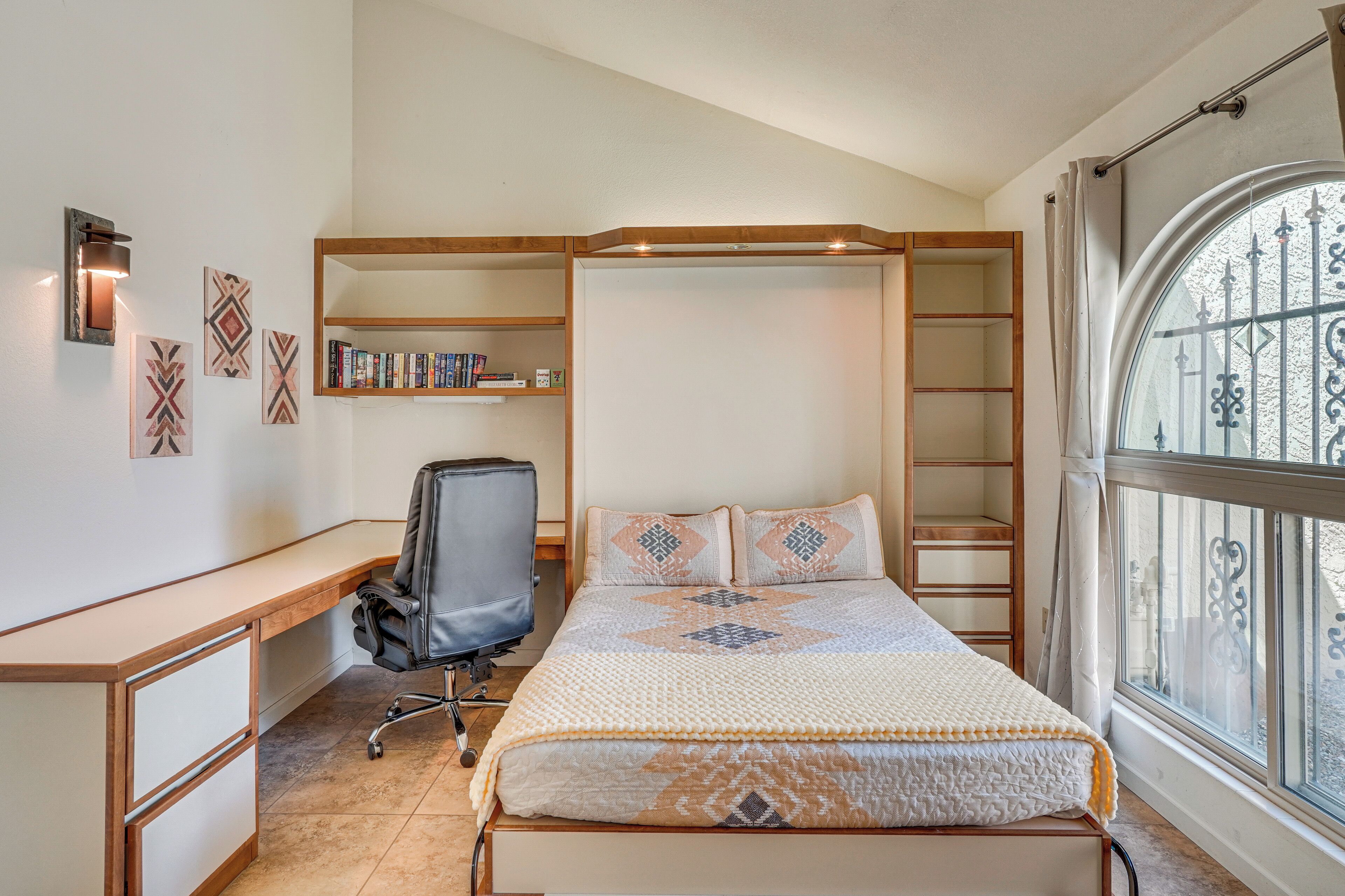 A bedroom with a bed, a desk, and a chair. There is a window with a view of the outside.