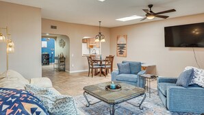 A living room with a sofa, a coffee table, and a TV mounted on the wall. There is a dining area with a table and chairs in the background. - Quiet Townhome w/ Mountain Views in Green Valley (Green Valley)