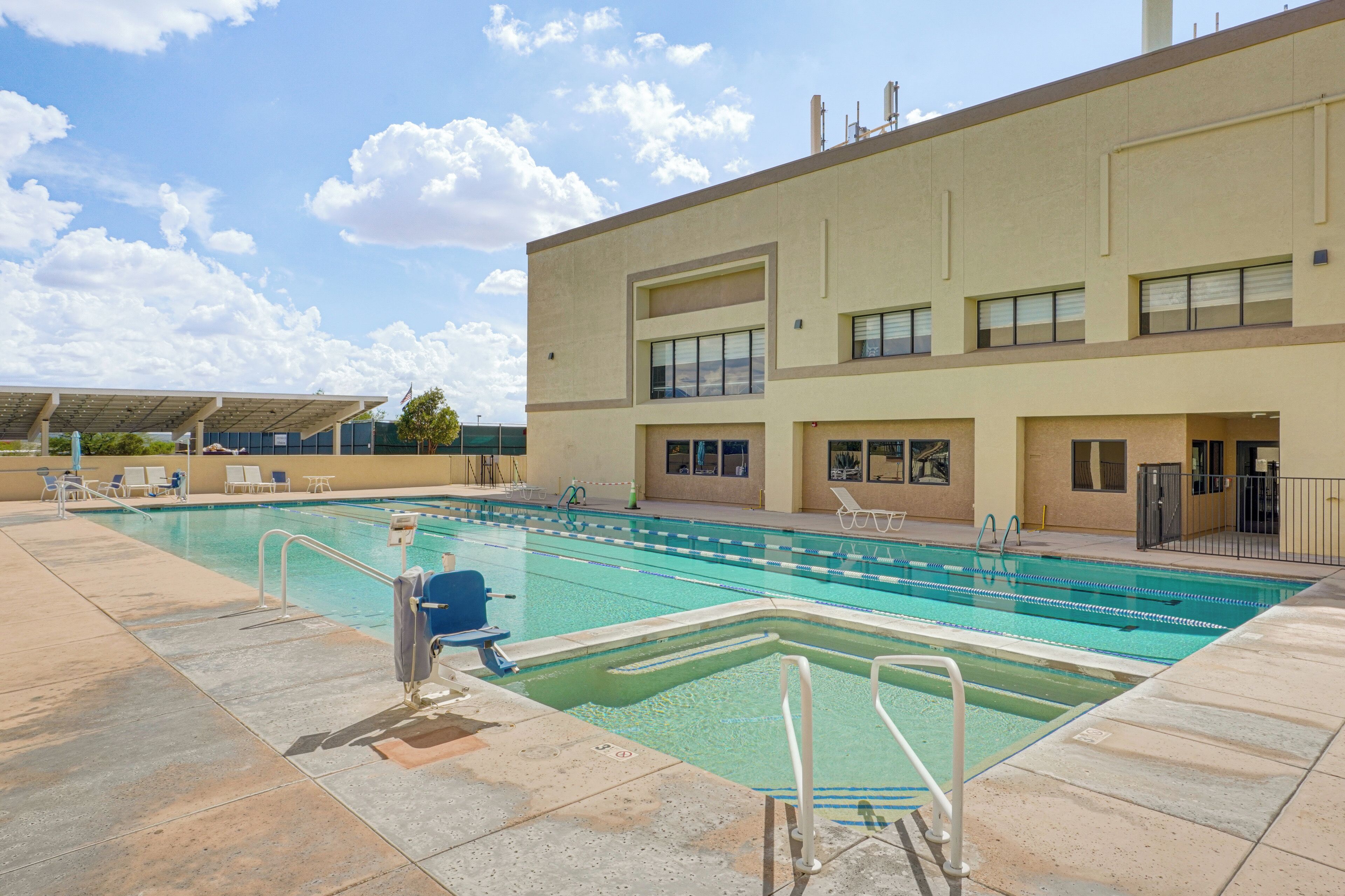 A swimming pool area with a building in the background, a covered seating area, and a clear sky.