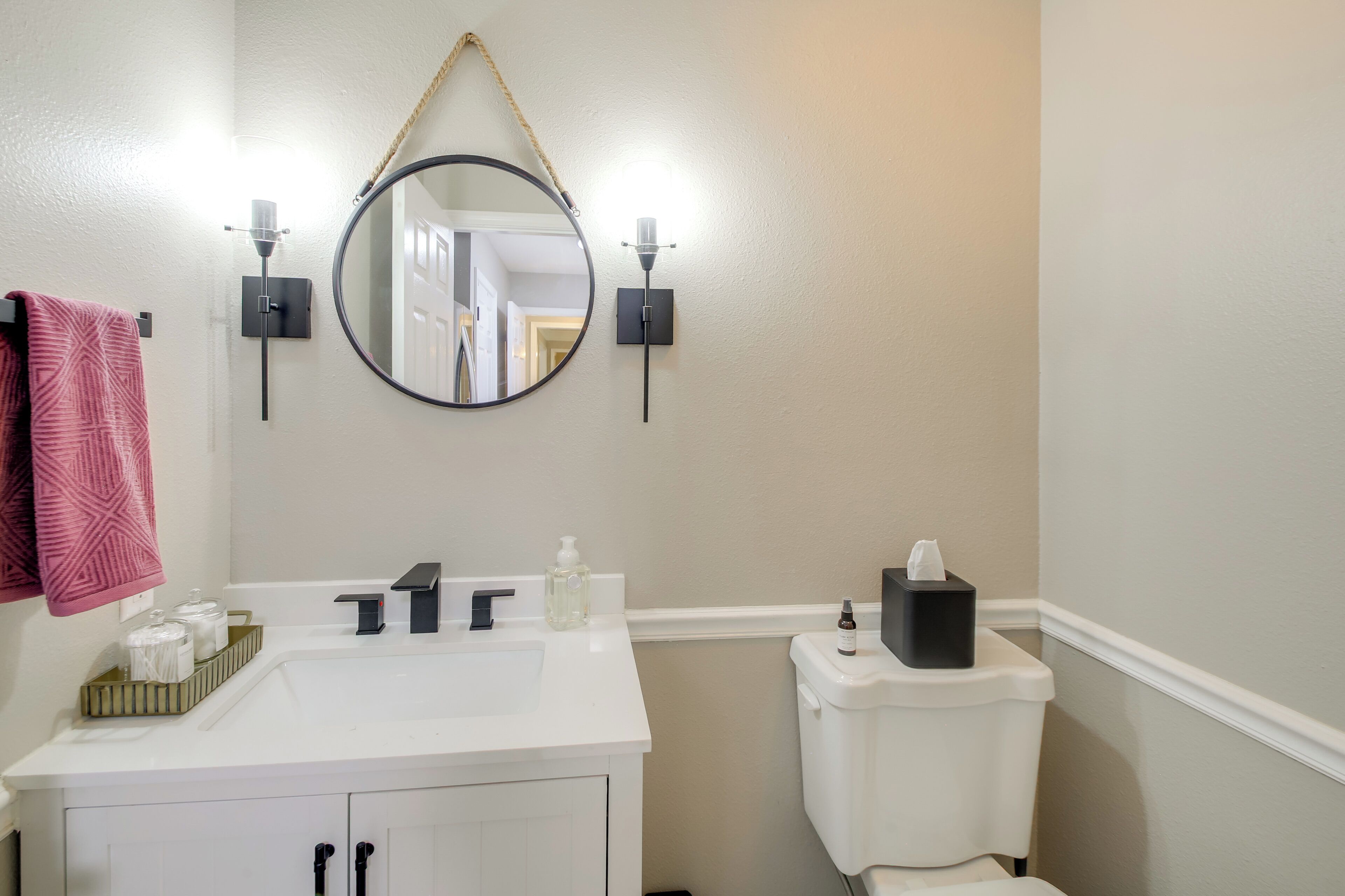 Un bagno con un lavandino bianco, uno specchio rotondo, due luci a parete, un water e un asciugamano rosa a sinistra.