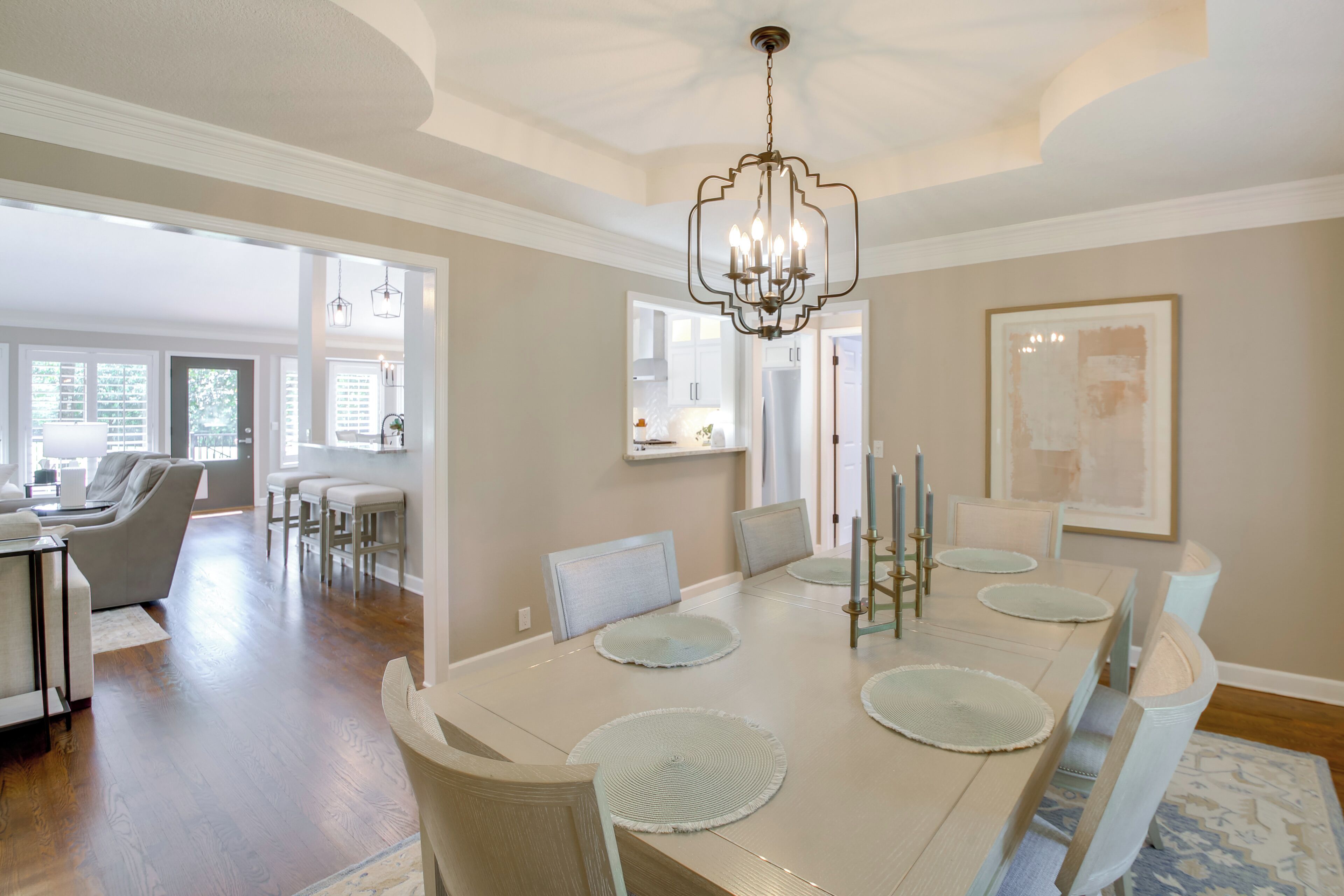 A dining room with a long table, chairs, and a chandelier. There is a kitchen in the background.