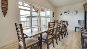 A dining area with a long wooden table and chairs, large windows, and a wooden shelf with decorative items.