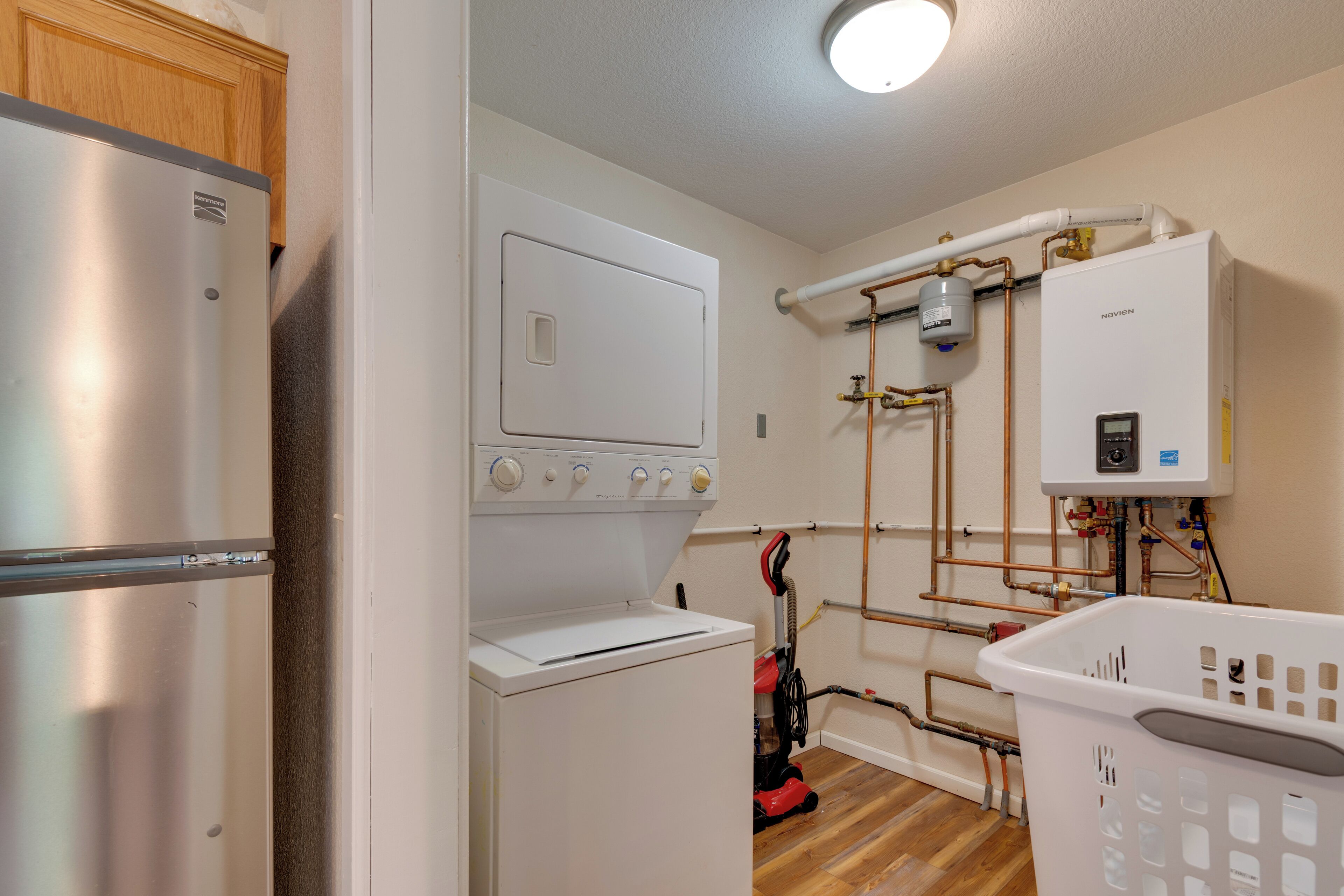 A compact laundry room with a stainless steel refrigerator, a washing machine, a water heater, and a red vacuum cleaner.