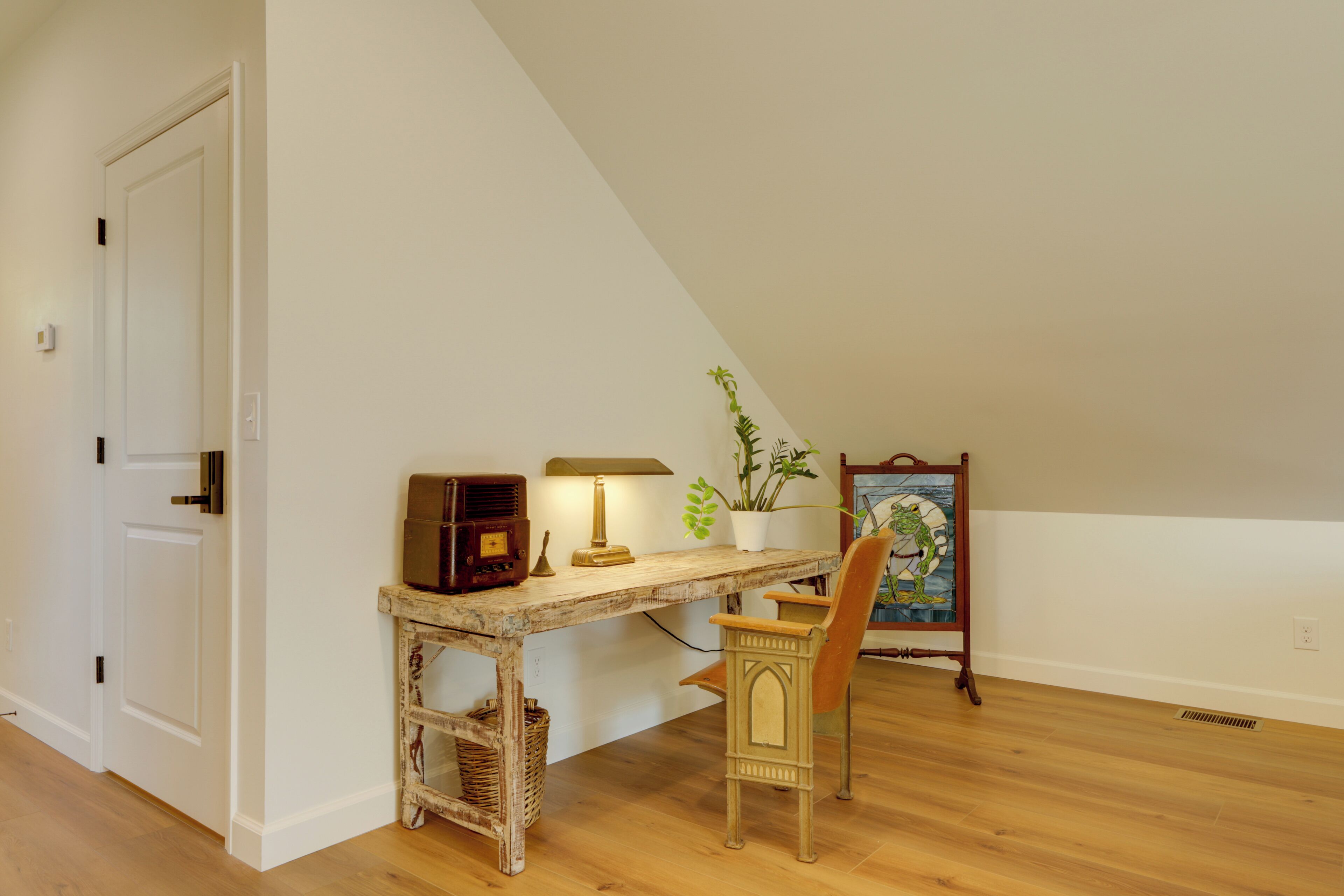 Um quarto com escrivaninha de madeira, uma cadeira, luminária, rádio, planta em vaso e um quadro na parede.